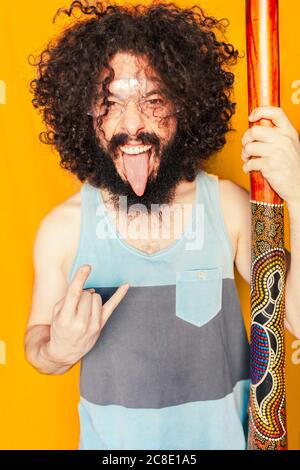 Verrückter Mann mit lockigen Haaren hält Didgeridoo beim Stehen heraus Zunge vor gelbem Hintergrund Stockfoto