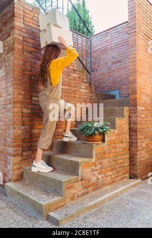 Junge Frau trägt Kisten auf dem Kopf, während sie über Treppen geht Durch Ziegelwand Stockfoto
