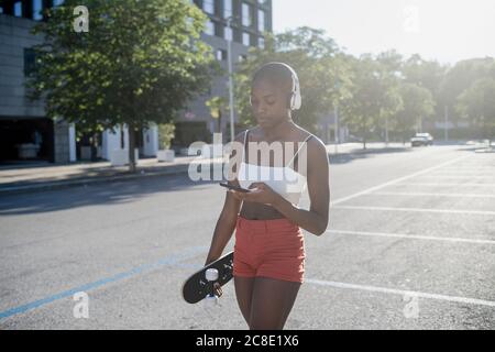 Junge Frau, die Musik über Kopfhörer mit dem Mobiltelefon hört, während Zu Fuß auf der Straße Stockfoto