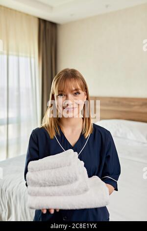 Lächelndes Zimmermädchen hielt Handtücher, während sie im Schlafzimmer im Hotel stand Stockfoto