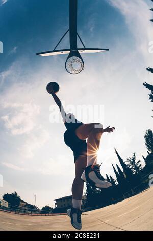 Low-Angle-Ansicht der weiblichen Sportlerin springen, während Dunking Basketball Gegen den Himmel Stockfoto