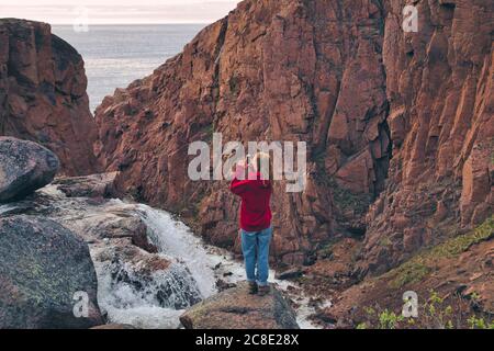 Junge Frau fotografiert Wasserfall, während sie auf felsigen Berg in Teriberka, Murmansk Oblast, Russland Stockfoto