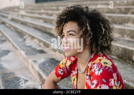 Nachdenkliche junge Frau lächelt, während sie auf der Treppe bei Sonnenschein sitzt Tag Stockfoto