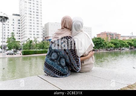 Muslimische Schwestern sitzen Arm um auf Stützmauer am Fluss In der Stadt Stockfoto