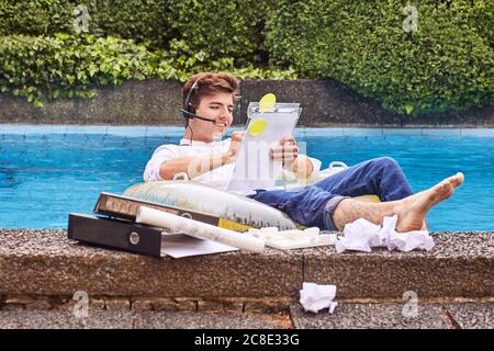 Junger Mann sitzt auf Luftmatratz im Schwimmbad und arbeitet Stockfoto