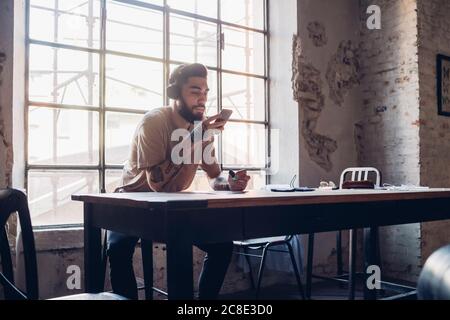 Kreativer junger Geschäftsmann mit Kopfhörer und Smartphone am Tisch sitzen Im Loft-Büro Stockfoto