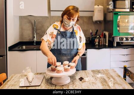 Bäckerin trägt Maske Dekoration Cupcakes auf Cakestand in der Werkstatt Stockfoto