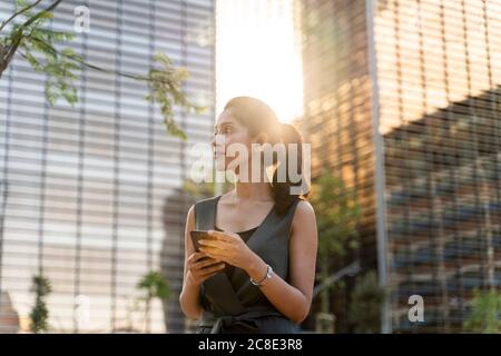 Durchdachte Geschäftsfrau hält Smartphone, während sie gegen moderne Gebäude steht Stockfoto