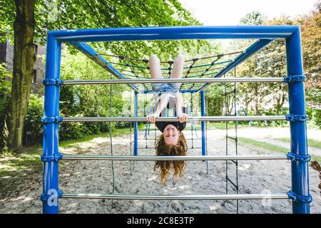 Lächelndes Mädchen, das auf dem Kopf auf dem Dschungel-Fitnessstudio auf dem Spielplatz hängt Im Park Stockfoto