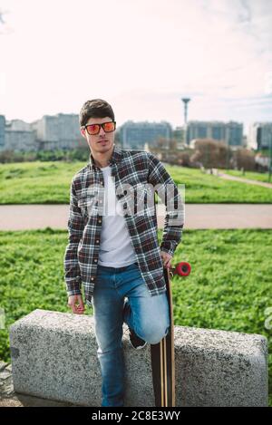 Mann mit Sonnenbrille hält Skateboard und steht gegen den Himmel Stadt Stockfoto