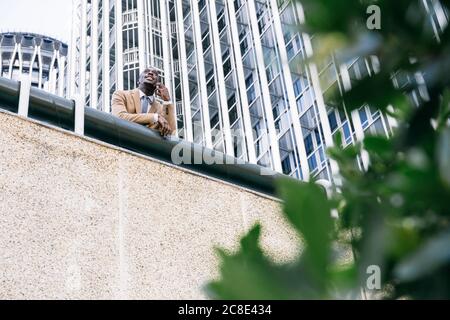 Junger Geschäftsmann, der sich auf ein Geländer in der Stadt stützt und spricht Am Telefon Stockfoto