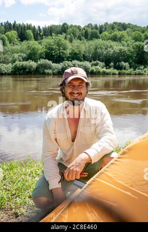Lächelnder Mann mit Mütze, der Zelt am See auf dem Campingplatz installiert Im Wald Stockfoto