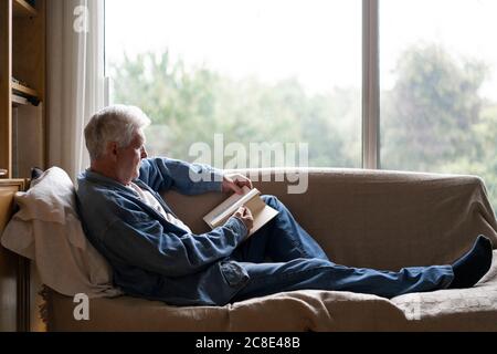 Ein älterer Mann liest ein Buch und entspannt sich auf dem Sofa am Fenster Zu Hause Stockfoto