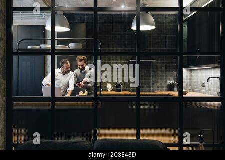Reifer Mann arbeitet auf Laptop in der Küche, Freund Blick auf Smartphone Stockfoto