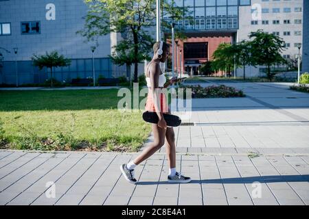 Junge Frau hält Skateboard mit Smartphone beim Gehen auf Fußweg in der Stadt Stockfoto