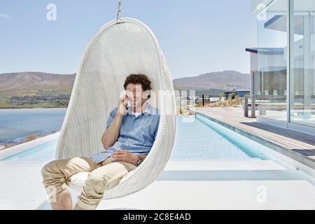 Mann, der in einem Hängesessel über dem Pool saß und sich unterhielt Das Telefon Stockfoto