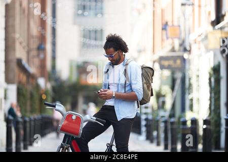Junger Mann mit Leihrad und Rucksack mit Handy in der Stadt, London, Großbritannien Stockfoto