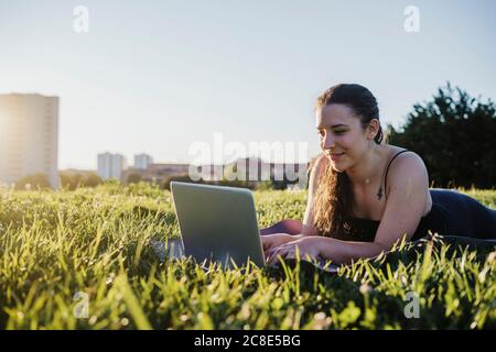 Sportliche Frau mit Laptop, während sie auf Gras in der Stadt liegt parken Sie an sonnigen Tagen Stockfoto
