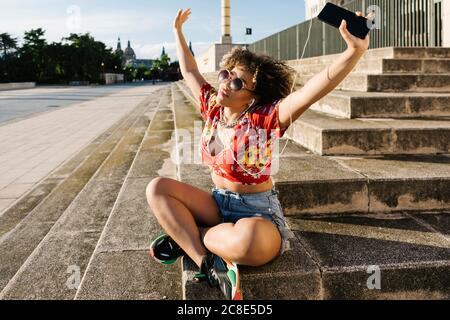 Trendige Frau, die Musik hört, während sie mit erhobenen Armen sitzt Nur wenige Schritte in der Stadt Stockfoto