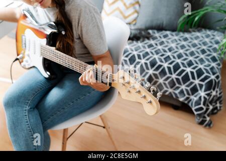 Frau spielt E-Gitarre, während sie zu Hause auf einem Stuhl sitzt Stockfoto