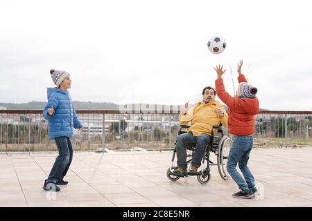 Mann sitzt auf Rollstuhl werfen Ball zu spielerischen Söhnen in parken Stockfoto