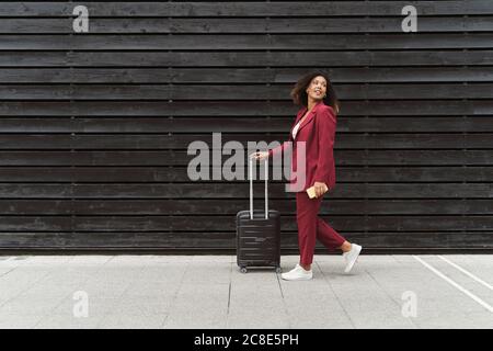 Junge Geschäftsfrau schaut zurück, während sie mit Gepäck auf dem Fußweg läuft Durch schwarze Holzmauer in der Stadt Stockfoto