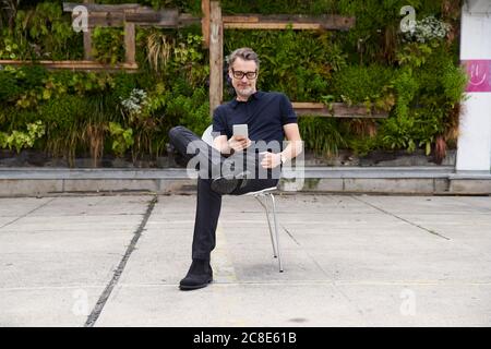 Reifer Mann mit Smartphone, während auf Stuhl sitzen Hof Stockfoto
