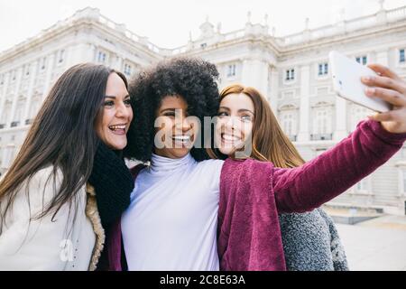 Glückliche junge Frau, die mit Freundinnen Selfie macht, während sie gegen den Königspalast von Madrid, Spanien, steht Stockfoto