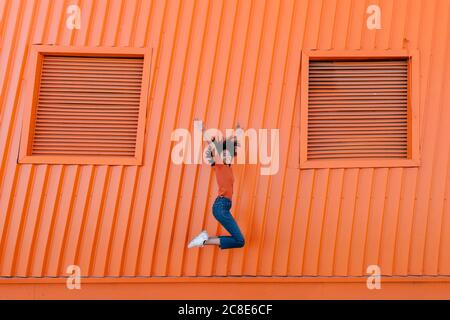 Fröhliche junge Frau, die mit den Armen gegen die orange Wand springen Stockfoto