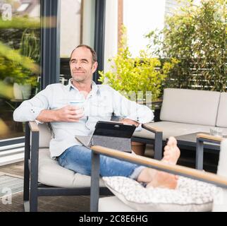 Geschäftsmann sitzt mit digitalem Tablet und Kaffeetasse auf einem Stuhl Im Hinterhof Stockfoto
