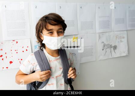 Junge mit Maske, die gegen Papiere steht, die an der Wand hängen Schule Stockfoto