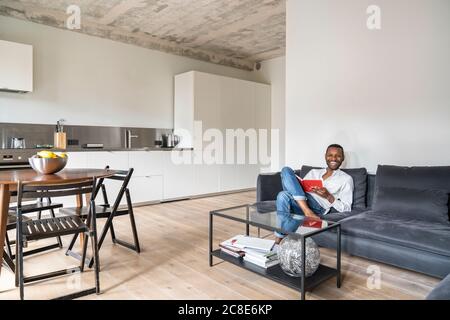 Lachender Mann mit Buch sitzt auf der Couch in der modernen Wohnung