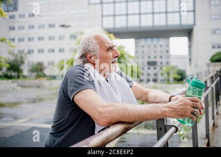 Älterer Mann hält eine Wasserflasche und hört Musik, während er sich im Stehen aufhält Geländer Stockfoto