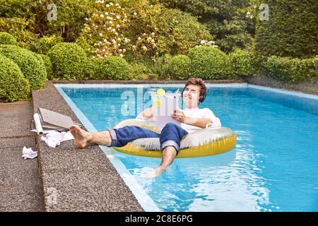 Junger Mann sitzt auf Luftmatratz im Schwimmbad und arbeitet Stockfoto