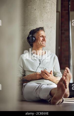 Lächelnder älterer Mann mit Kopfhörern, der in einem Loft Musik hört Flach Stockfoto