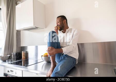 Lachender Mann, der zu Hause auf der Küchentheke sitzt und isst apfel Stockfoto