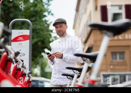 Lächelnder Mann, der sich am Parkplatz ein Fahrrad über ein Smartphone ausgeliehen hat In der Stadt Stockfoto