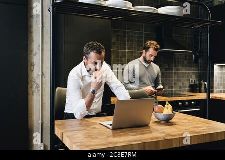 Reifer Mann arbeitet auf Laptop in kitchen Apfel, Freund Blick auf Smartphone Stockfoto