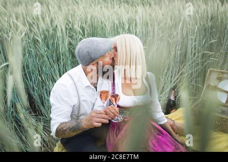 Pärchen hält Sektgläser küssend, während sie inmitten von Pflanzen sitzt Land Stockfoto