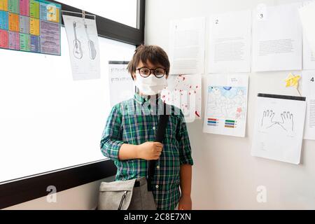Junge trägt Maske mit Tasche, die an Wand und Fenster steht In der Schule Stockfoto
