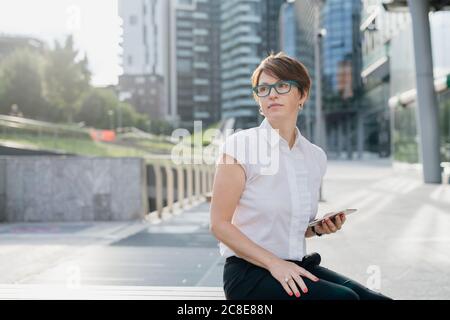 Durchdachte Geschäftsfrau hält Smartphone, während sie gegen Gebäude sitzt Stadt Stockfoto