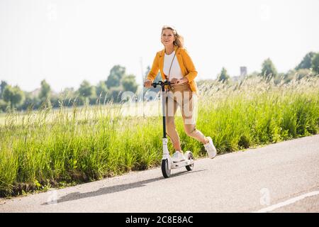 Spielerisch reife Frau Reiten Elektro-Push-Roller auf der Straße während Sommer Stockfoto