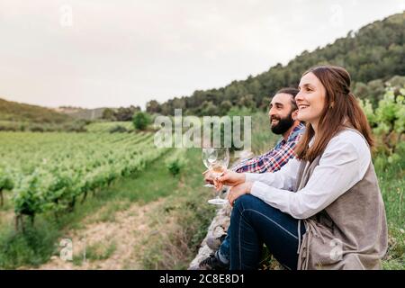 Glückliches Paar hält Weißweingläser, während es am Weinberg sitzt Stockfoto