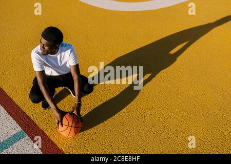 Nachdenklicher junger Mann hält Basketball hocken auf Sportplatz während Sonniger Tag Stockfoto