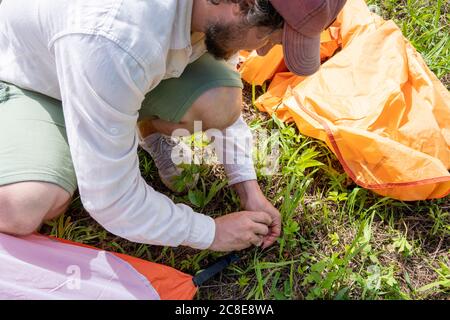 Mittelerwachsener Mann mit Kappe, der Zelt auf Gras aufstellend Auf dem Campingplatz Stockfoto