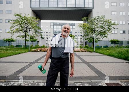 Glücklicher älterer Mann, der Wasserflasche hält, während er auf dem Fußweg steht Gegen Gebäude Stockfoto