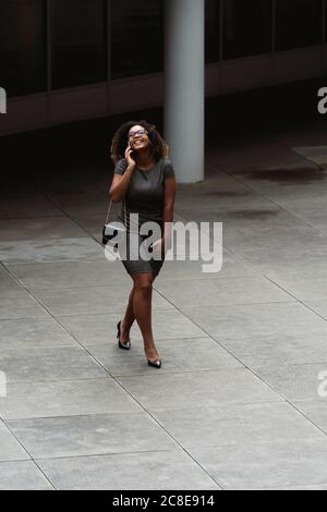 Fröhliche junge Geschäftsfrau im Gespräch über Smartphone, während zu Fuß auf Fußweg in der Stadt Stockfoto
