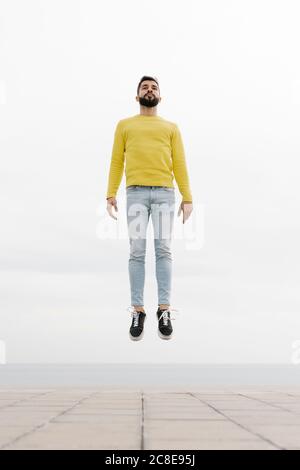 Sorgloser bärtiger junger Mann, der auf dem Fußweg gegen die weiße Wand springt Stockfoto