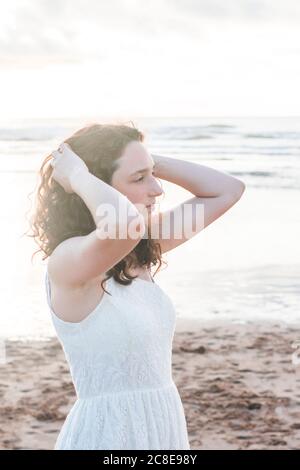 Nachdenklich junge Frau trägt weißes Kleid mit Händen im Haar Stehen am Strand Stockfoto