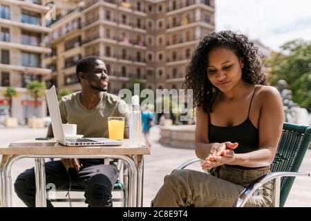 Junge Frau reinigt die Hände mit Desinfektionsmittel, während der Mann sitzt Straßencafé Stockfoto